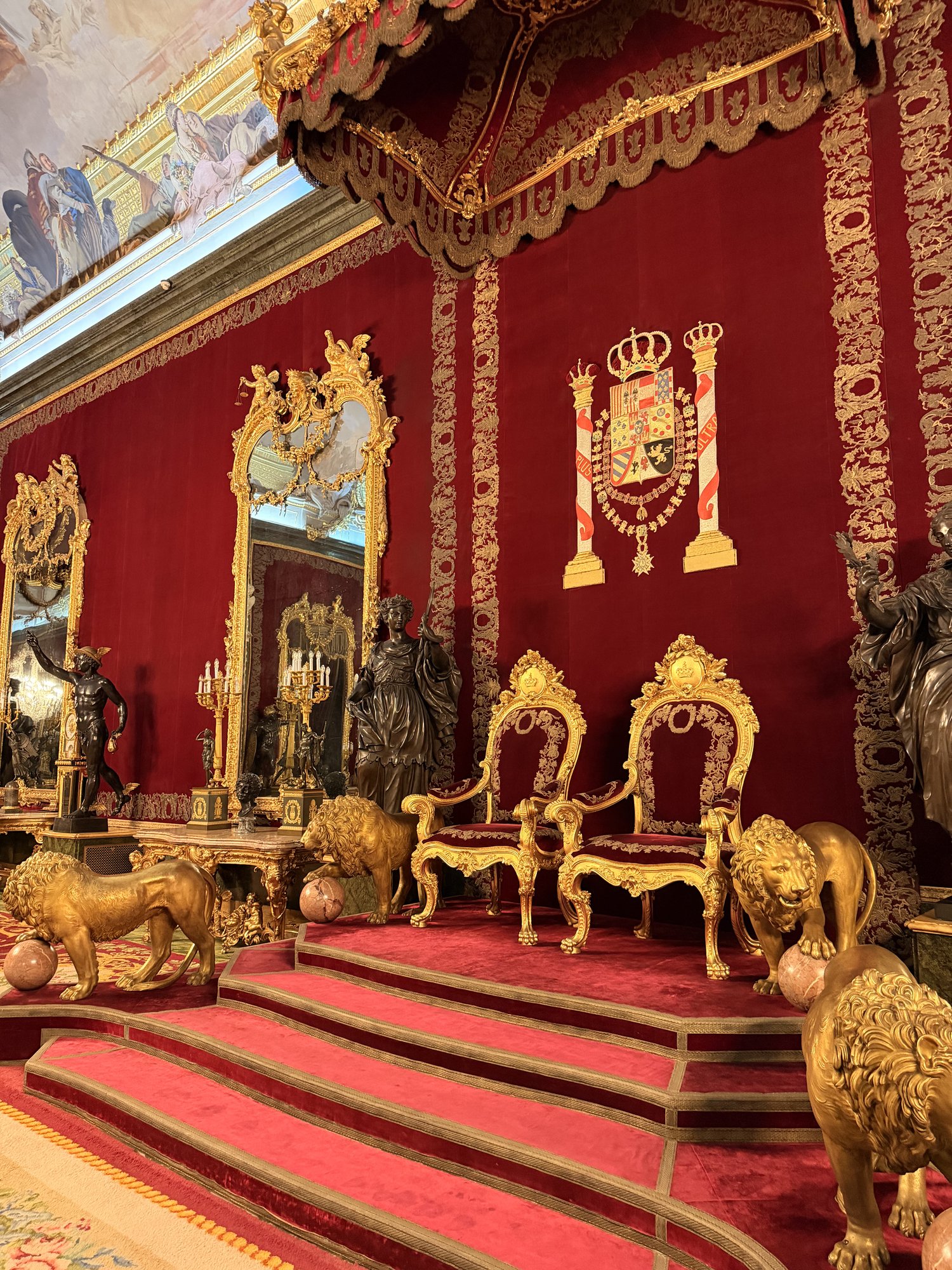 The Throne Room of the Royal Palace of Madrid with red velvet walls, gilded twin thrones, golden lions, and a painted ceiling fresco